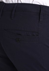 Donkerblauwe broeken met een gladde textuur, een achterzak met een zwarte knop en subtiele stikseldetails langs de naden.