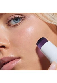 Vit cylindrisk hudvårdsprodukt med ett lila lock, hålls nära kinden. Matt hudtextur och klar hy är synlig.