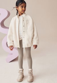 Veste en sherpa couleur crème avec des boutons-pression, haut blanc, leggings côtelés gris clair et bottes beiges à lacets avec une bordure en fourrure.