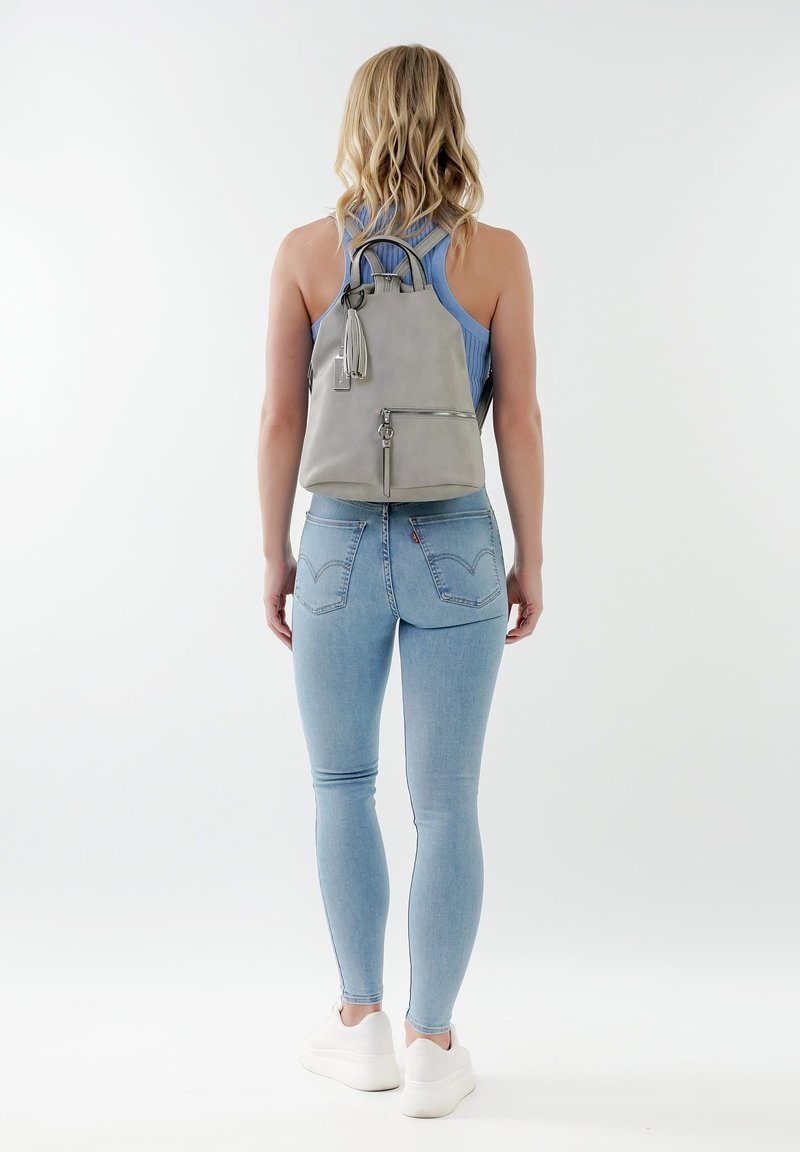 Zaino in pelle grigio con doppi manici superiori, scomparto frontale con zip e decorazione a frange. Il modello indossa una maglietta senza maniche blu e jeans chiari.