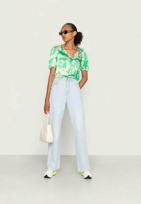 Mulher vestindo uma blusa floral verde, jeans de perna larga azul-claro, tênis brancos, óculos de sol escuros, segurando uma bolsa branca, de pé contra um fundo simples.