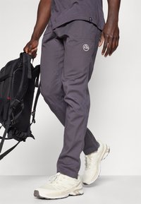 Γκρίζα εξωτερικά παντελόνια με εφαρμοστή σχεδίαση, που διαθέτουν πλάγιες τσέπες και λογότυπο, συνδυασμένα με αθλητικά παπούτσια ανοιχτού χρώματος.