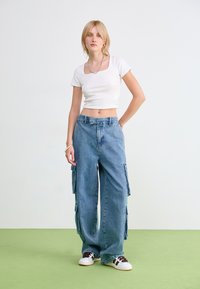 T-shirt cropped blanc, jean cargo bleu à jambes larges avec poches latérales et bords effilochés, complété par des baskets blanches avec des accents foncés.