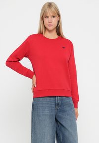 Felpa rossa con scollo rotondo e polsini a coste, con un piccolo logo a forma di cuore sul petto. Indossata con jeans in denim larghi.