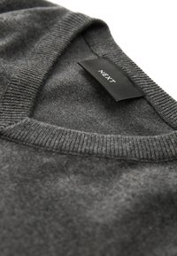 Pull en tricot gris foncé avec encolure en V et ourlet côtelé. L'étiquette indique "NEXT." Le tissu présente une texture lisse avec de légères variations de teinte.