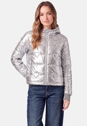 Frau trägt silberne, metallische, gesteppte Jacke mit Kapuze und Herzmustern, kombiniert mit blauen Jeans, stehend vor weißem Hintergrund.