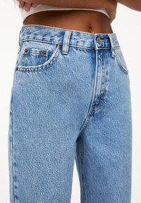 Torse habillé d'un jean denim bleu clair taille haute avec bouton et passants pour ceinture, les mains reposant juste au-dessus de la taille.