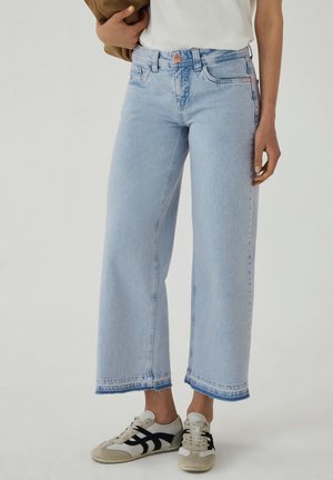 Person trägt hellblaue, weit geschnittene Cropped-Jeans und weiße Sneakers mit schwarzen Akzenten, steht vor einem einfarbigen Hintergrund.