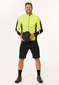 Neon gul cykeljacka med svarta detaljer, dragkedja fram, ihop med svarta shorts, håller en svart cykelhjälm. Svarta cykelskor.