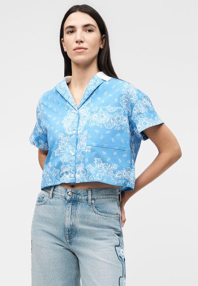 Γυναίκα με κοντομάνικο μπλε πουκάμισο με παγιέτα σε μοτίβο paisley και ανοιχτό μπλε τζιν με κεντητά πλαϊνά patches, τα χέρια στηρίζονται στη μέση.