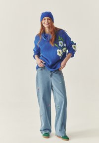 Pulover tricotat albastru cu broderie florală pe mâneci, asociat cu blugi largi de culoare albastră deschisă, având mici accente brodate colorate. Încălțări verzi.