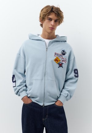 Mladý muž so svetlohnedými vlasmi, oblečený v svetlomodrej mikine na zips s nášivkami s témou baseballu a tmavomodrých džínsoch, ruky v vreckách.