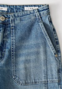 Jean en denim bleu clair en gros plan montrant le bouton, la fermeture éclair, les poches avant et latérales avec des coutures visibles et la texture du tissu.