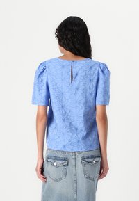 Blouse bleu clair avec des manches courtes bouffantes, texture florale et une ouverture en goutte d'eau à l'arrière, associée à une jupe en denim bleu clair.
