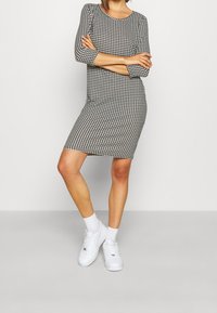 Černobílé šaty s houndstooth vzorem, ke kolenům, kulatý výstřih, tříčtvrteční rukávy, spárované s bílými teniskami a ponožkami.