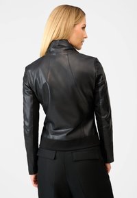 Kvinde med blond hår iført en tætsiddende sort læderjakke og sorte bukser, vist fra ryggen mod en ensfarvet baggrund.
