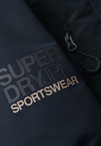 Zwarte sportkleding stof met reliëf "SUPER DRY JPN" logo in zwart en goud. Kenmerkt zich door een ritsluiting en een gestructureerd oppervlak.