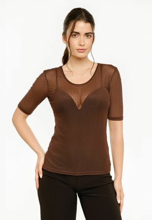 Frau mit dunklen Haaren im Pferdeschwanz trägt ein braunes, durchsichtiges gepunktetes Oberteil über einem passenden Camisole und schwarzen Hosen, posiert mit einer Hand auf der Hüfte.