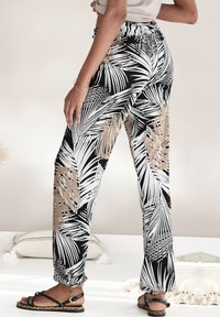 Zwart-wit palmblad patroon broek met een elastische tailleband en gathers bij de enkels. De stof heeft een gladde textuur.