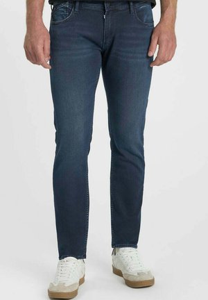 Mann trägt dunkelblaue Slim-Fit-Jeans und weiße Sneakers, steht vor einem schlichten Hintergrund.