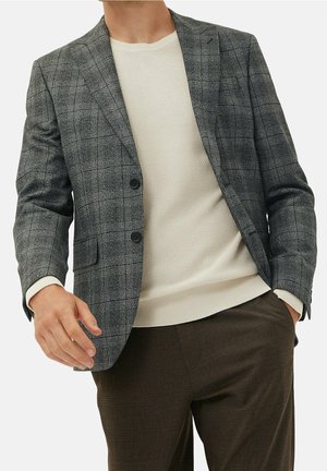 Homme portant un blazer à carreaux gris, un pull en maille crème et un pantalon à carreaux brun foncé, une main dans la poche sur un fond uni.