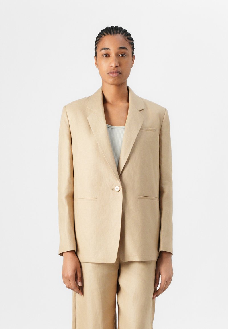 Veste de costume en lin beige avec une fermeture à un bouton, des revers crantés et des poches avant. Texture lisse, coupe ajustée et design minimaliste.