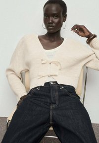 Cardigan testurizzato color crema con due fiocchi, indossato sopra una maglietta bianca, abbinato a jeans scuri a vita alta. Modella seduta su una sedia.