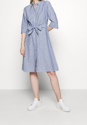 Woman in knee-length, light blue, button-down dress with three-quarter sleeves and tied waist belt, wearing white platform sneakers.