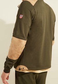 Chaqueta verde oliva con detalles de ante beige en los hombros y las mangas. Presenta un cordón en el dobladillo y un parche con el logo en la manga.