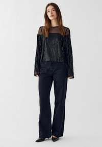 Czarna bluzka z długimi, frędzlami i przezroczym dekoltem, ozdobiona metalowymi niciami. Połączona z ciemnymi, szerokimi dżinsami i czarnymi butami.