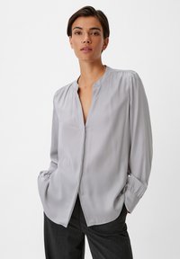 Hellgraue Bluse aus glattem Stoff mit V-Ausschnitt, gerafften Schultern und Knopfverschluss. Lange Ärmel mit Manschetten.