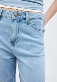 Κοντινό πλάνο ανοιχτόχρωμων τζιν denim με εμφανές κουμπί μάρκας Lee, τσέπη και θηλιά για ζώνη, φορεμένα με ανοιχτόχρωμο κοντό τοπ.