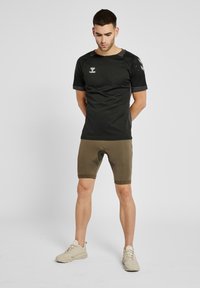 Schwarzes kurzärmliges Sportshirt mit Mesh-Akzenten; kombiniert mit olivgrünen enganliegenden Shorts und neutralfarbigen Sportschuhen.