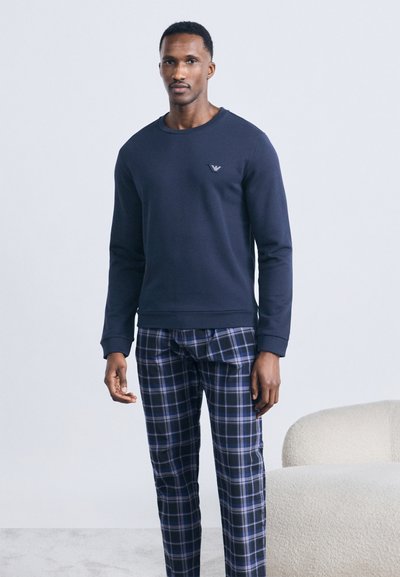 Άνδρας που στέκεται φορώντας μπλε μπλούζα με μικρό λογότυπο και μπλε καρό πιτζάμες μπροστά σε απλό φόντο.