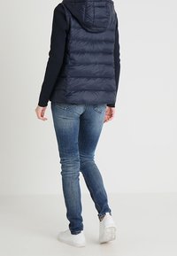 Tmavě modrá vatovaná vesta s kapucí, s prošívanou texturou a předními švy, nošená přes černé tričko s dlouhým rukávem, modré džíny a bílé tenisky.