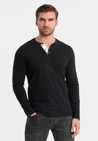 Camisa henley de manga larga negra con un tapón de botones blanco y marrón, hecha de tela suave; combinada con jeans oscuros.