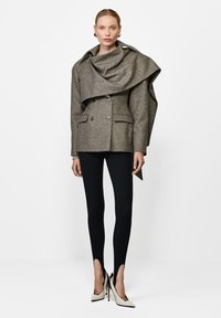 Szary wełniany płaszcz dwurzędowy z luźnym kołnierzem i dwiema przednimi kieszeniami, zestawiony z czarnymi legginsami i dwukolorowymi szpilkami w kształcie stożka.