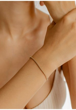 Brazalete de oro delgado con un diseño suave y flexible, que presenta un delicado patrón de cadena y un pequeño broche, llevado en una muñeca.
