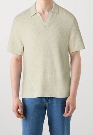 Mann trägt ein strukturiertes beige Polohemd mit kurzen Ärmeln, kombiniert mit blauen Jeans, vor einem schlichten Hintergrund.