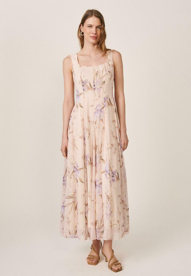 Mujer con vestido sin mangas color crema hasta los tobillos con estampado floral morado y sandalias de tacón color tostado, de pie frente a un fondo beige liso.