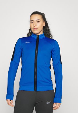 Giacca da track Nike blu con pannelli laterali neri, colletto alto, maniche lunghe e zip frontale. Texture liscia, design aderente, logo sul petto.