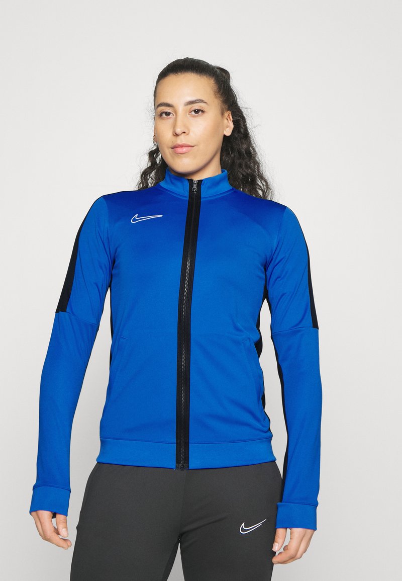 Blauwe Nike trainingsjack met zwarte zijpanelen, hoge kraag, lange mouwen en rits aan de voorkant. Soepele textuur, aansluitend design, logo op de borst.