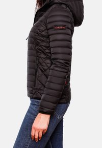 Žena v černé prošívané bundě s kapucí a červeným logem "MARIKOO" na rukávu, kombinující s modrými džíny, na jednobarevném bílém pozadí.