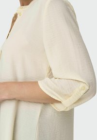 Cream-colored shirt with a textured fabric. Features a round collar, buttoned placket, and rolled-up sleeves secured with buttons.