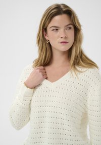 Jeune femme aux cheveux blonds portant un pull blanc à col en V en maille, des boucles d'oreilles en or en forme de créoles et un bracelet en or, regardant sur le côté.