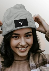 Gray knit beanie with a ribbed texture and a black patch featuring a white deer head design. The hat is snugly fitted to the head.