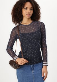 Marineblauw mesh longsleeve top met contrasterende strepen bij de manchetten, met een subtiel patroon en een aansluitend ontwerp. Bruine schoudertas in de hand.