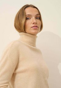 Beżowy sweter z golfem wykonany z miękkiego materiału, z prążkowanymi detalami przy dekolcie i gładką teksturą. Model zaprezentowany na jasnym tle.