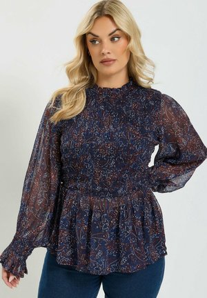 Blonde vrouw die een donkerblauw patroon blouse met lange mouwen en geplooide details draagt en een blauwe spijkerbroek, poseert met één hand op de heup.