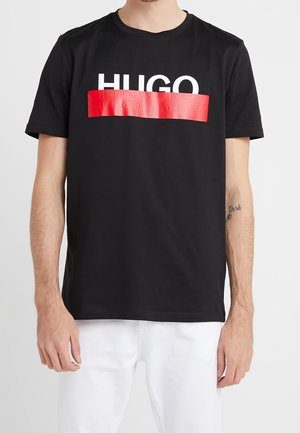 Mężczyzna w czarnej koszulce z krótkim rękawem z białym tekstem "HUGO" częściowo zakrytym czerwonym błyszczącym paskiem na klatce piersiowej, w połączeniu z białymi spodniami.